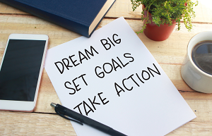 Cell phone, book, coffee and notepad reading "Dream Big, Set Goals, Take Action" on a desk.
